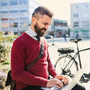 Hipster businessman commuter with bicycle on the way to work in city, sitting on a bench and using laptop.
