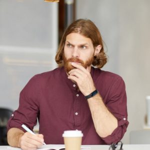 Red haired bearded businessman sitting at the table making notes and looking pensively with woman in the background in cafe