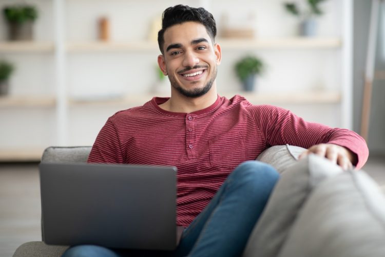 Smiling arab guy reclining on sofa with laptop at home, copy space. Handsome young middle-eastern man enjoying weekend, using notebook, watching moovie or surfing on dating website
